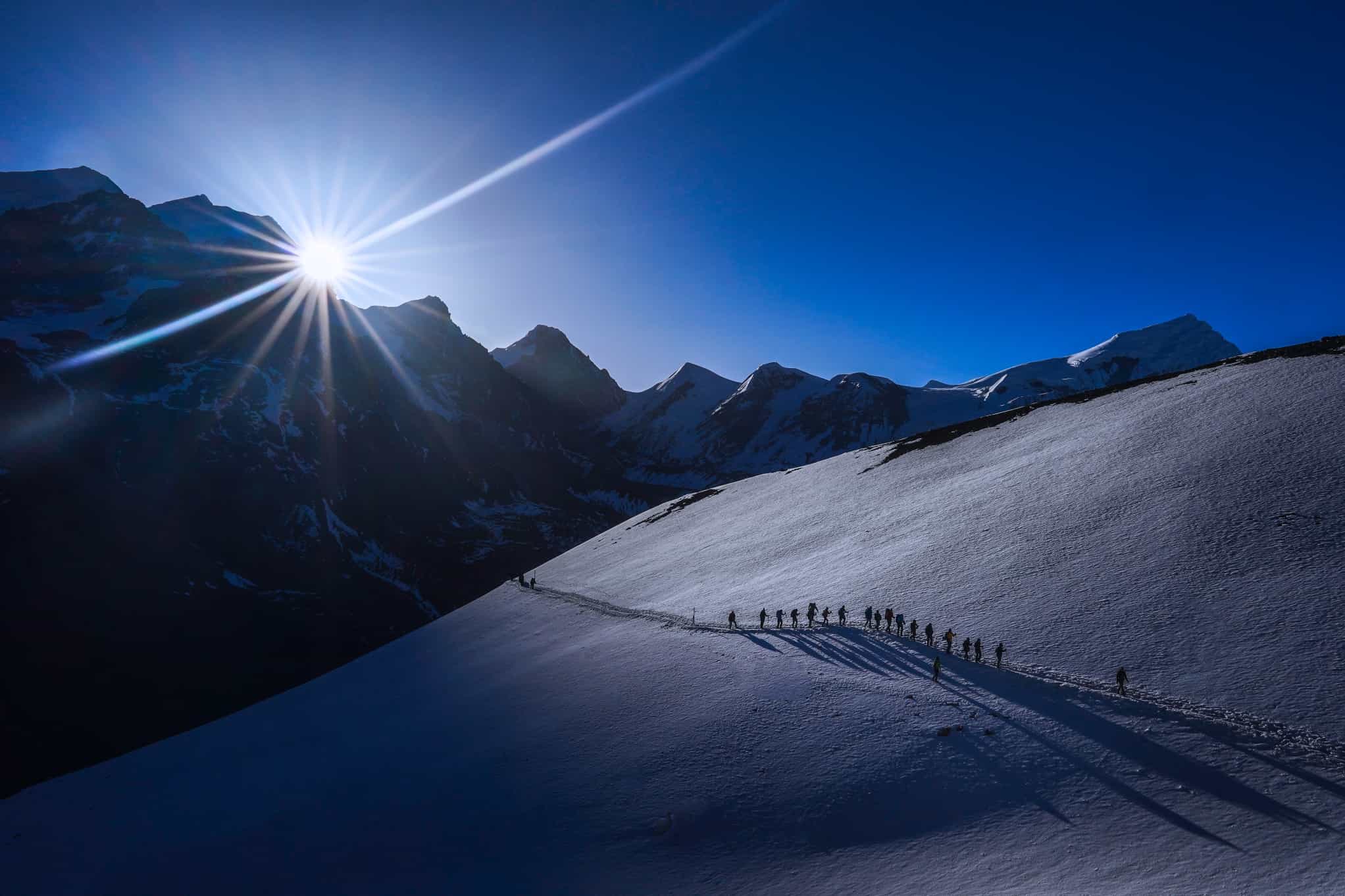 A string of trekkers on the Thorong La Pass, with the sun rising over the mountains in the background, Nepal.