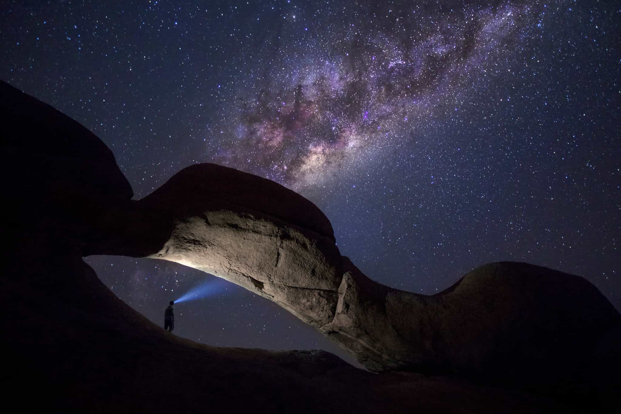 Hiker with a head torch standing in a rock arch near Spitzkoppe at night with the Milky Way visible above.